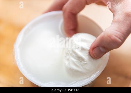 Macro primo piano di tenuta di mano con burrata fresca mozzarella formaggio italiano in contenitore di plastica acquistato in magazzino con salamoia e colore bianco Foto Stock