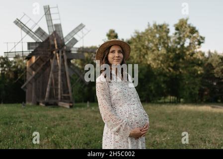 Un colpo medio lungo di una donna incinta nello sfondo del mulino a vento che sta tenendo il suo ventre con le mani sulla fattoria. Una gravidanza felice. Una signora in un Foto Stock