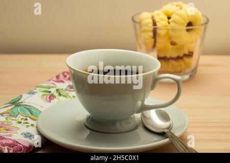 Una tazza di caffè e una ciotola di vetro con biscotti di burteria e un tovagliolo a motivi floreali su una superficie di legno. Foto Stock