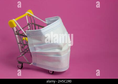 Un carrello in un supermercato, con una maschera medica protettiva contro il coronavirus, su sfondo rosa. Il concetto di protezione contro il covid Foto Stock