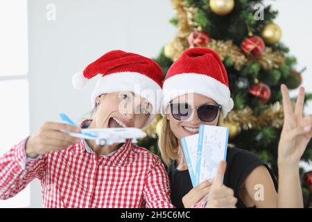 Ritratto di due giovani donne in babbo natale cappelli in possesso di aereo e biglietti contro di albero di Capodanno Foto Stock