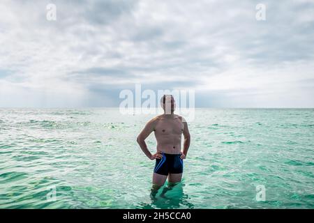 Giovane muscoloso fit felice uomo adulto in spiaggia in piedi nuoto in chiaro trasparente turchese oceano acqua di mare con orizzonte oceano sfondo nuvoloso giorno Foto Stock