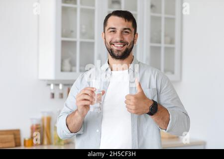 Sorridente bell'uomo caucasico adulto tenere il vetro con acqua e mostrare il pollice in su Foto Stock