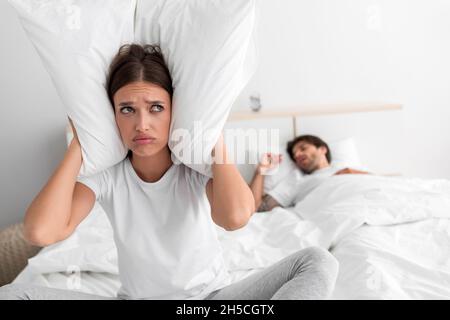 Despaired la moglie europea millenaria siede sul letto copre le orecchie con il cuscino e soffre di russare del marito dormiente Foto Stock