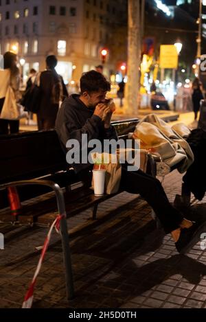 Uomo senza casa in abiti casual seduti su panca e mangiare hamburger da asporto di notte sulla strada affollata della città Foto Stock