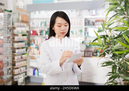Donna cinese cercando medicina con notebook vicino scaffali in farmacia Foto Stock