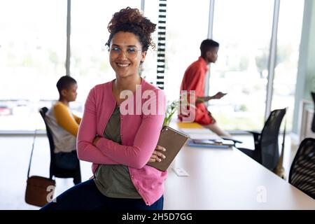 Ritratto di sorridente donna d'affari biraciale in casuali seduti con le braccia incrociate in ufficio creativo Foto Stock