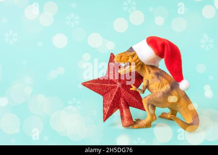 Il Rex del dinosauro in cappello rosso di Babbo Natale tiene la stella nelle relative zampe su sfondo blu la vigilia di Capodanno o la vigilia di Natale scheda di festa di arte creativa idea per Merry Foto Stock