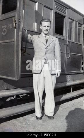 1940, storico, un uomo in piedi su una piattaforma ferroviaria che tiene la porta di una carrozza ferroviaria aperta, Inghilterra, Regno Unito. La porta è contrassegnata CON LA TERZA classe, indicando che è la terza classe di carrozza o pullman, dietro LA SECONDA e LA PRIMA classe. Anche se molte delle compagnie ferroviarie avevano abolito la seconda classe da quest'epoca, molti dei treni che da e per Londra avevano mantenuto la diversa classe di carrozze. Alla fine degli anni '50, tuttavia, la terza classe era stata rinominata seconda classe e poi molto più tardi negli anni '80, divenne nota come classe 'standard'. Foto Stock