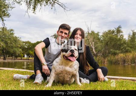 Due persone sedute accanto ad un cane bully americano in un parco vicino ad un fiume Foto Stock