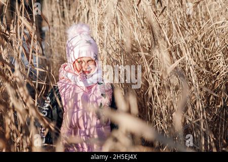 Bambina in erba secca. Bambino in rosa caldo vestito inverno esplora il thicket di canne in giorno di sole nella natura e gode di passeggiata Foto Stock