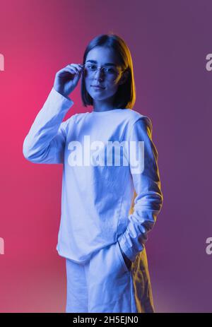 Primo piano ritratto di giovane ragazza carina, studente in abiti casual ascoltando musica isolato su rosa-viola studio backgroud in neon. Foto Stock