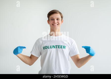 Molto orgoglioso di essere un volontario. Giovane ragazzo in guanti medici che punta alla sua uniforme, sorridendo a macchina fotografica sopra la parete di luce Foto Stock