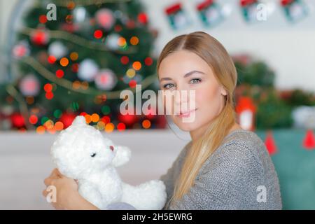 Bella donna con carino piccolo orsacchiotto bianco sul divano vicino decorato albero di Natale. Bella e tranquilla serata di Natale a casa. Foto Stock