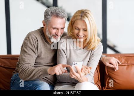 La coppia anziana felice con lo smartphone sul divano trascorre tempo libero in social network, scorrimento feed, guardare video, uomo grigio-capelli sta mostrando smth a sorridente bella donna Foto Stock