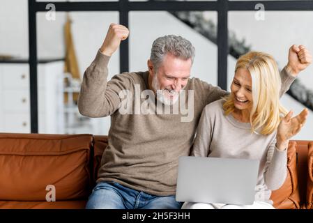 La coppia anziana utilizza un computer portatile per guardare partite sportive o lotterie online seduti su un comodo divano nel soggiorno. Allegri sposi di mezza età che alzano le mani in su nel gesto di vittoria, trionfo Foto Stock