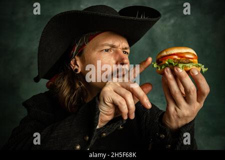 Ritratto di un uomo brutale, pirata medeival toccare hamburger isolato su sfondo scuro. Foto Stock