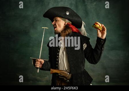 Ritratto di un uomo brutale, pirata medeival tenendo hamburger isolato su sfondo scuro. Foto Stock