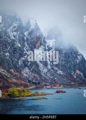 Una tipica scena paesaggistica norvegese, con montagne di granito innevate e ricoperte di nebbia che sovrastano una minuscola casa rossa, isolata in un fiordo blu chiaro. Foto Stock