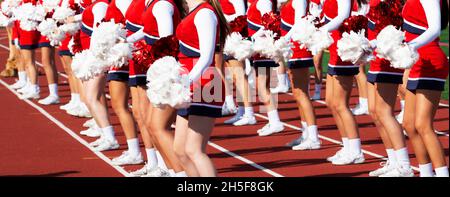 I cheerleader delle scuole superiori indossano uniformi rosse e bianche che si acclamano ai margini di una partita di calcio Foto Stock