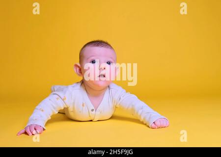 Bambino ragazzo guarda sorpreso mentre si trova sul suo pancino, studio sfondo giallo. Primo piano bambino di quattro mesi Foto Stock