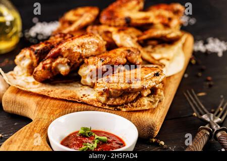 Primo piano delle cosce di pollo alla griglia. Appetitose carni di pollame alla griglia, salsa e forchette su un tagliere di legno. Sfondo rustico scuro. Foto Stock