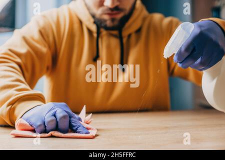 Primo piano ritratto delle mani dell'uomo con guanti di gomma che puliscono tavolo di legno. Uomo che fa un po 'di lavoro di pulizia in casa. Igiene a casa. Foto Stock