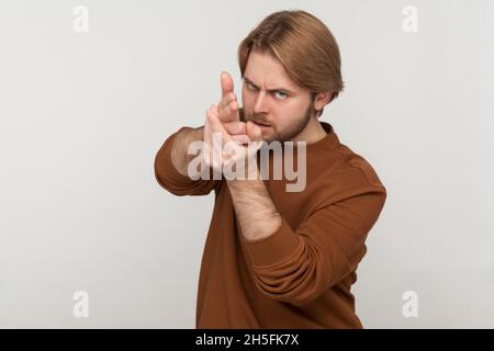 Ritratto di pericoloso giovane adulto bel uomo con barba indossare felpa, puntando con pistole a mano alla macchina fotografica, fingendo di sparare. Studio interno girato isolato su sfondo grigio. Foto Stock