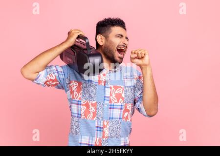 Giovane adulto bearded pazzo uomo che tiene il registratore a nastro sopra la spalla, ascoltare musica, cantare con microfono cospicuo, indossare camicia blu. Studio interno girato isolato su sfondo rosa. Foto Stock