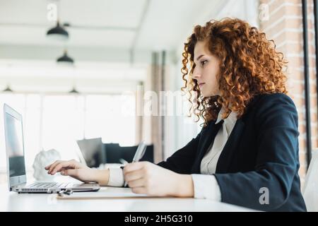 Bella giovane donna con capelli rossi in un vestito da lavoro lavora in un ufficio con un computer portatile Foto Stock
