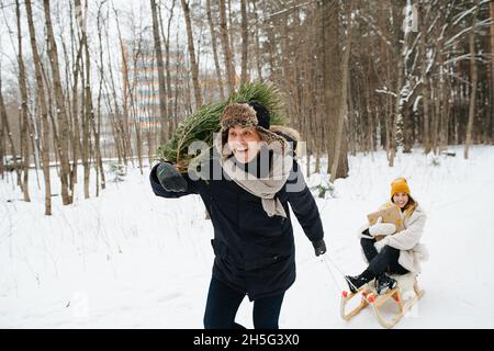 Una coppia felice. Un uomo con un albero di Natale sulla spalla corre attraverso il parco invernale e tira una slitta con una donna dietro di lui. Foto Stock