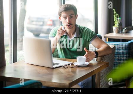 Ritratto di uomo d'affari che indossa una T-shirt verde, lavora su un laptop, punta un dito, indica la direzione della fotocamera e guarda infastidito, facendo la scelta. Foto al coperto vicino alla grande finestra, sfondo del caffè. Foto Stock