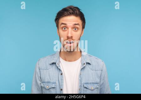 Ritratto di uomo stupito indossando una camicia in denim, in piedi con la bocca aperta di sorpresa, ha sconvolto l'espressione, sente notizie incredibili. Studio interno girato isolato su sfondo blu. Foto Stock