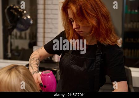 Primo piano. Parrucchiera femminile di colore rosso medio-invecchiato con le braccia tatuate in una stretta uniforme nera pettinando, raddrizzando ed asciugando i capelli biondi di Foto Stock