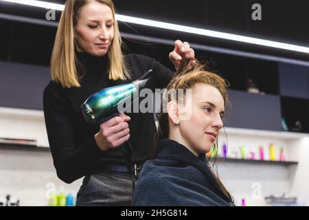 Master donna parrucchiere asciuga i capelli della ragazza con un asciugacapelli dopo il lavaggio in un salone di bellezza. Foto Stock