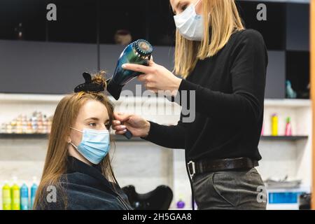 Master donna parrucchiere in una maschera medica asciuga i capelli della ragazza con un asciugacapelli e pettini dopo il lavaggio in un salone di bellezza. Covid-19 pandemia e. Foto Stock