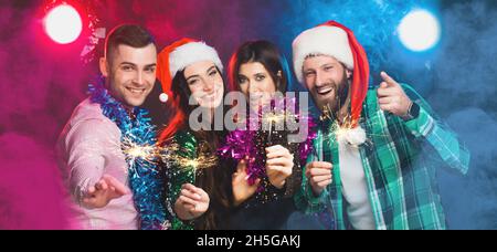 Festa di Capodanno con i migliori amici. Un gruppo di giovani con cappelli di Capodanno e scintillanti in mano sta guardando la fotocamera Foto Stock