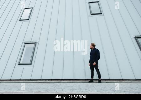 Ingegnere maschile, architetto maschile si erge sullo sfondo di una fabbrica o costruzione, tubi e edificio grigio, background tecnologico, urbano moderno Foto Stock