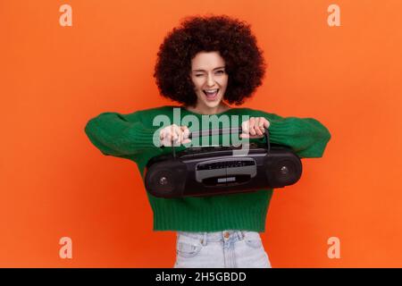 Divertente donna eccitata con acconciatura Afro indossando maglione verde casual stile regge registratore a nastro, premendo il pulsante, ascoltando musica ad alto volume. Studio interno girato isolato su sfondo arancione. Foto Stock