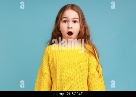 Ritratto di scioccato stupito bambina guarda la macchina fotografica a bocca aperta, avendo espressione sorpresa, indossando giallo casual stile maglione. Studio interno girato isolato su sfondo blu. Foto Stock