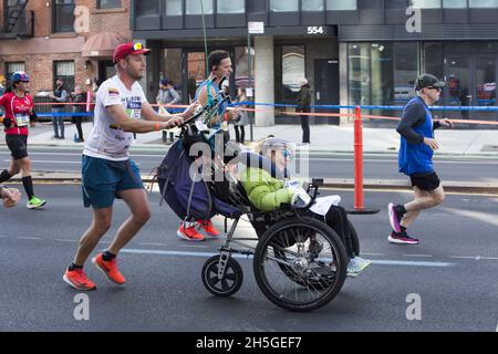 50° anniversario New York City Marathon Race nel 2021 dopo nessuna gara nel 2020 fare alla pandemia di Covid-19. L'uomo con una bandiera ecuadoriana sulla sua maglietta corre spingendo una donna paraplegica dall'aspetto felice davanti a lui sulla 4th Avenue attraverso la sezione Brooklyn della gara. Foto Stock