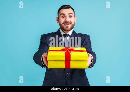 Ritratto di uomo d'affari sorridente bearded che indossa il vestito di stile ufficiale scuro che dà la scatola del regalo avvolta e sorridente alla macchina fotografica, festa felice. Studio interno girato isolato su sfondo blu. Foto Stock