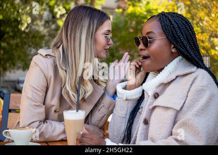 Due giovani amiche si gustano il caffè insieme, seduti a un tavolo, chiacchierando, sussurrando e gossipando. Concetto di diversità e tempo libero. Foto Stock