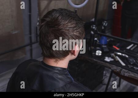 Un ragazzo senza trirned con capelli scuri siede su una sedia in un salone di fronte ai servizi e un taglio di capelli in un barbiere. Foto Stock