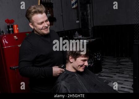 Un parrucchiere positivo o barbiere di aspetto europeo taglia un cliente soddisfatto con capelli scuri in un parrucchiere. Parrucchiere. Foto Stock