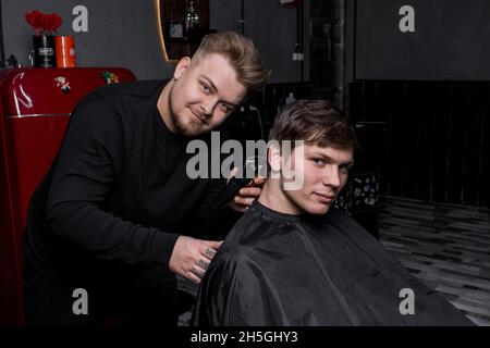 Un parrucchiere positivo o barbiere di aspetto europeo taglia un cliente soddisfatto con capelli scuri in un parrucchiere. Parrucchiere. Foto Stock