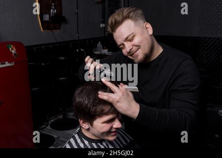 Un parrucchiere positivo o barbiere di aspetto europeo taglia un cliente soddisfatto con capelli scuri in un parrucchiere. Parrucchiere. Foto Stock