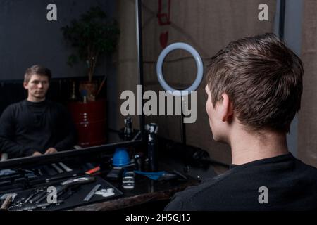 Un ragazzo timido con lunghi capelli scuri si siede in una sedia in un barbiere di fronte al riflesso nello specchio. La necessità di servizi di parrucchiere. Foto Stock