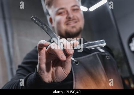 La mano di un barbiere o professionale può contenere un rasoio dritto utensile affilato per la rasatura e la cura della barba, primo piano. Foto Stock