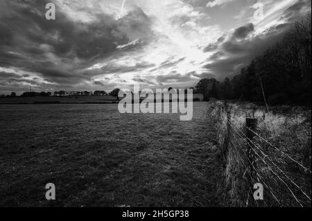 Immagine monocromatica ad alto contrasto in bianco e nero della recinzione del filo spinato in campo verde sotto il cielo nuvoloso con il sole sullo sfondo Foto Stock